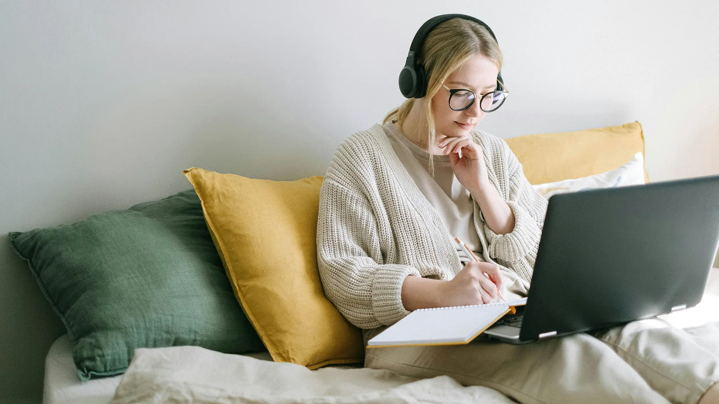 Photo of a relaxed looking woman reclining on sofa, with a laptop, taking notes while listening to headphones.